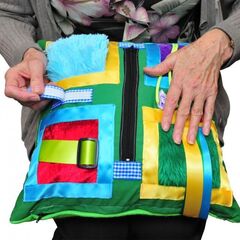 Elderly hands holding sensory cushion with tactile buttons, zips, and colorful materials
